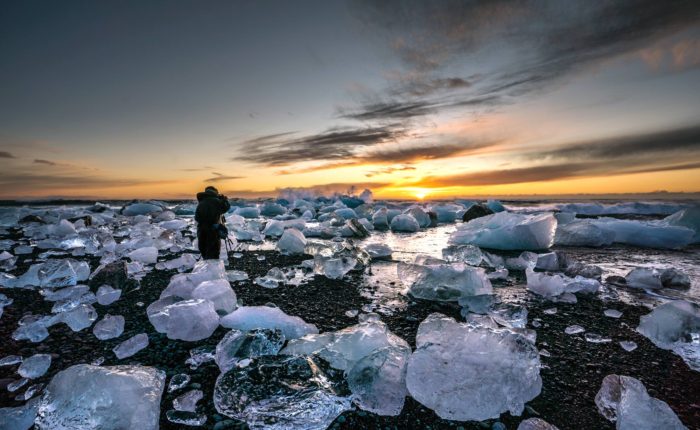 A man stands on a black sand beach surrounded by icebergs