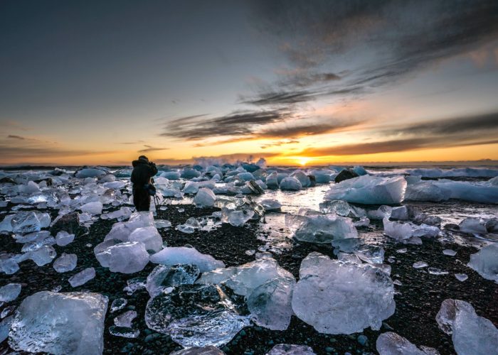 A man stands on a black sand beach surrounded by icebergs