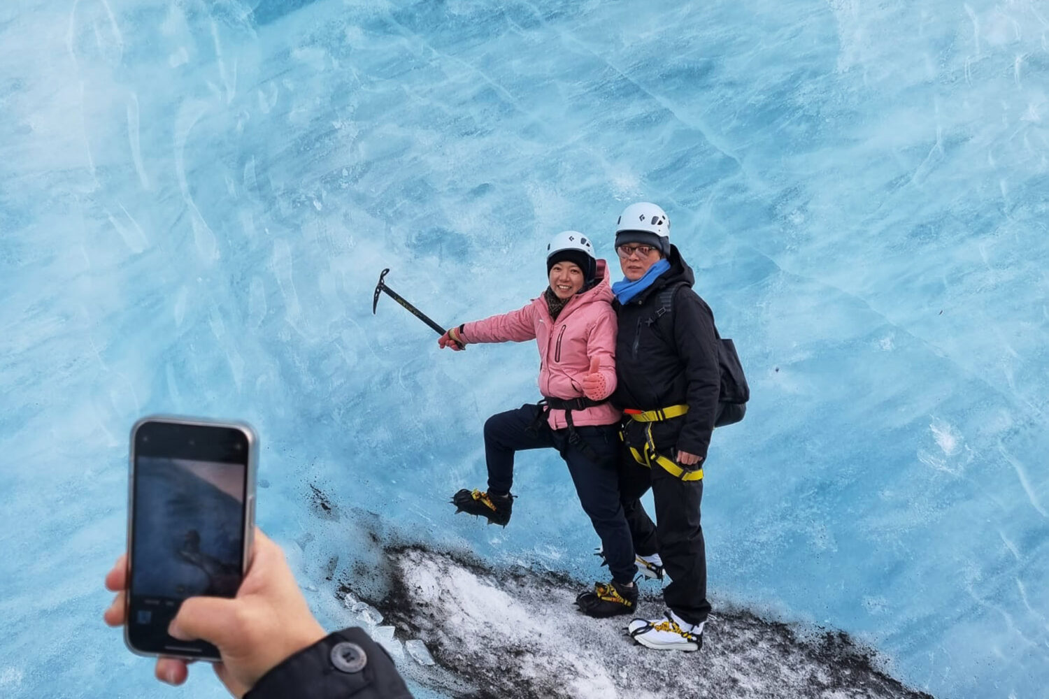 A person is taking a photo on it's phone of a couple standing by an ice wall on the glacier