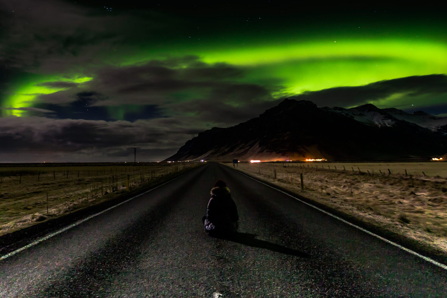 Girl sits on road in Iceland looking at the Northern Lights